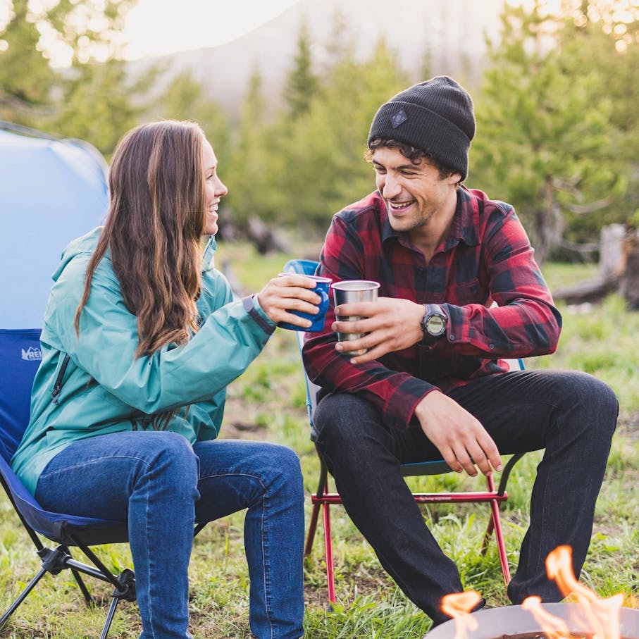 Man and woman sitting in camping chairs in front of a campfire in BD apparel with cups in their hands