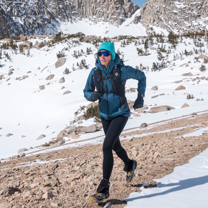 A runner treks across a trail with lofty snowy peaks behind her. 