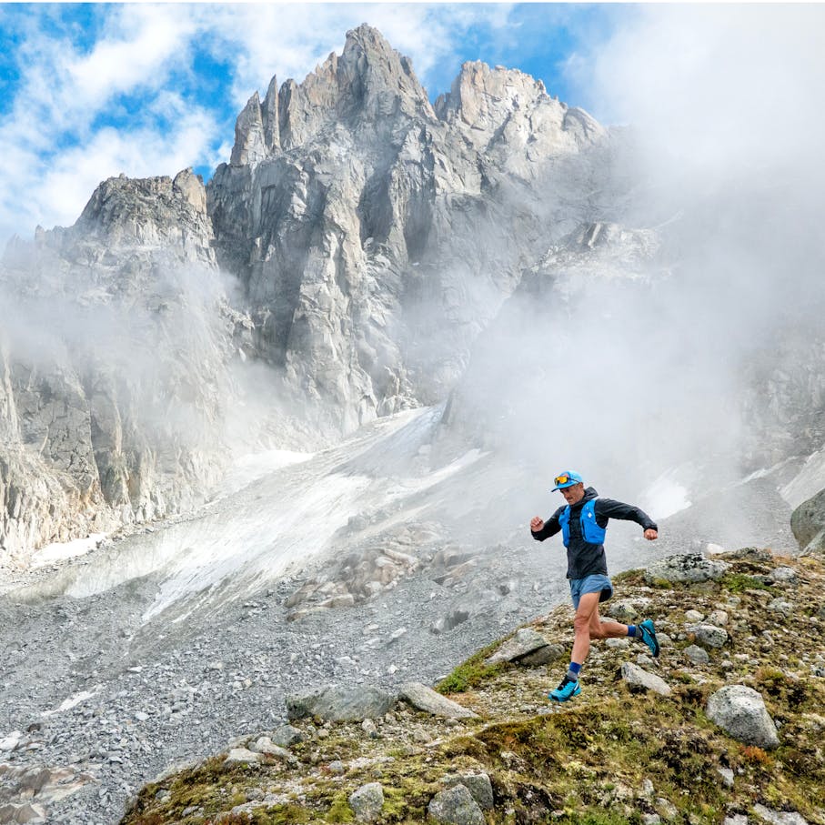 A runner on a grassy slope with lofty peaks behind him.